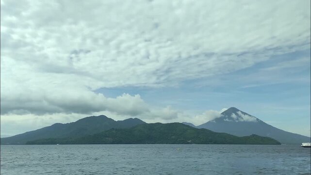 A cinematic drone timelapse captures the dynamic movement of altocumulus clouds over the iconic volcanic peaks and lush islands of North Maluku