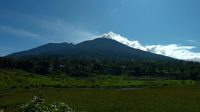 A dynamic ground-level timelapse captures white clouds moving swiftly behind the silhouetted volcanic peak of Mount Marapi in Padang, West Sumatra