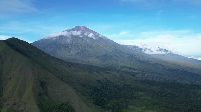 A cinematic drone shot pans slowly to the left, showcasing the majestic volcanic peak of Mount Rinjani and the surrounding green ridges of the Sembalun highlands