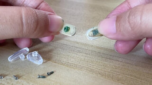 A macro shot of fingers holding and showing a pair of old, yellowed airbag nose pads with green oxidation buildup, next to new clear ones on a wooden surface.