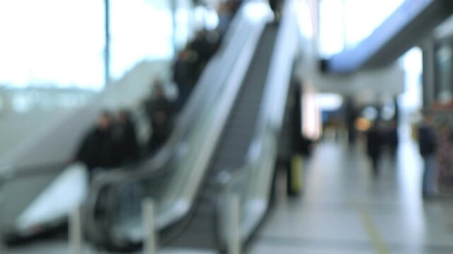 Blurred indoor escalator with people moving up and down. Background shows airport or mall hallway. Light reflects off polished floor and glass walls. No animals or plants visible