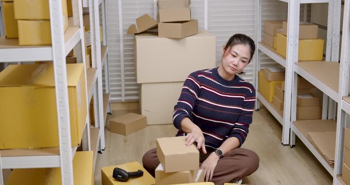 Asian woman small business owner sitting on warehouse floor, stacking parcel box carelessly, showing tired emotion, managing online shopping inventory alone inside home based e-commerce storage area