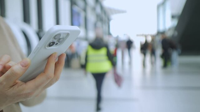 Person holds smartphone, focused on screen. Background shows blurred airport corridor. Travelers walk with luggage, out of focus. Scene suggests waiting or navigating transit