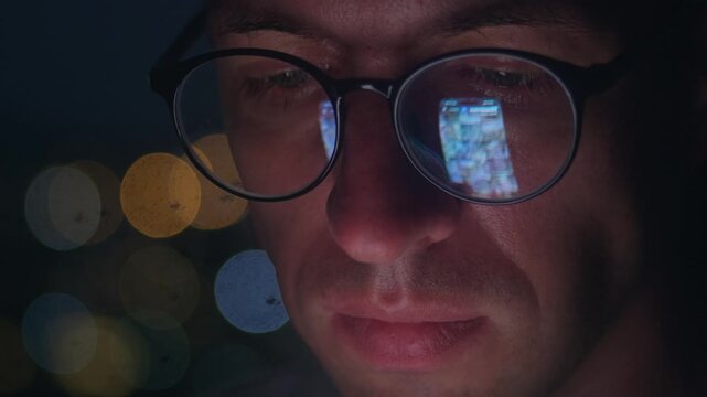 Extreme close up portrait side view man in glasses scrolling mobile phone at night city background, screen reflected in lenses. Focused millennial male face lit by mobile phone, social media browsing 