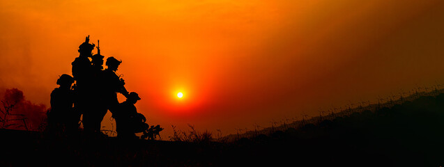 Silhouetted soldiers in formation review tactical equipment and rifles against a dramatic fiery sunset, wearing camouflage uniforms and maintaining a somber, professional stance. © AU USAnakul