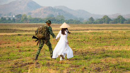 Obraz premium Soldiers and a Woman in White Dress Walking Across Grassy Field in Afghanistan Carrying Firearms and Backpacks, Creating a Somber Environment. A camouflage soldier and a woman wearing a white dres