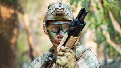 Obraz premium Southeast Asian Soldier Aiming Rifle in Forest, Wearing Camouflage Gear and Helmet, Displaying Focused Alertness and Readiness for Immediate Action in a Wooded Terrain, Military Training Scene