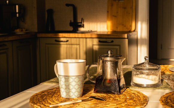 Cozy morning tea ritual: glass teapot, cup and sugar bowl on a sunlit kitchen table, warm golden-hour light and calm domestic charm. Tea time table setup for a cozy tea party at home