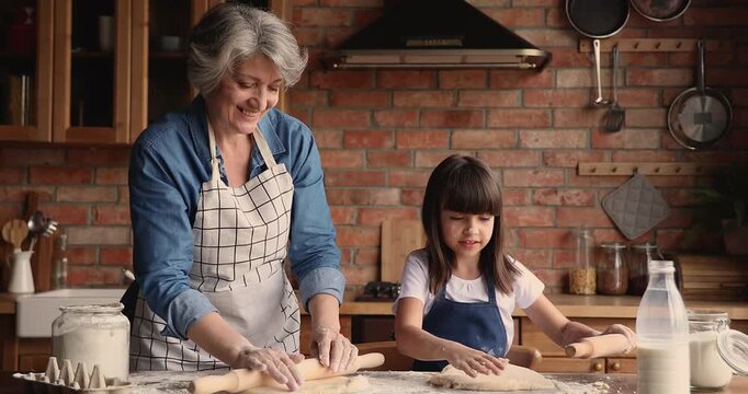 Loving grandmother teaching to cook child granddaughter, rolling dough with rolling-pins for homemade pastries or bread in domestic kitchen at home, multigenerational family baking together. Cooking