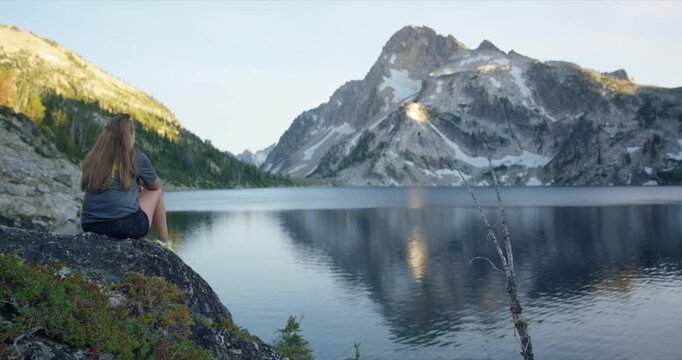 Blonde white woman sits on cliff edge overlooking Sawtooth Lake and rugged alpine mountain peak