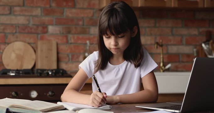 Schoolgirl sitting at kitchen table with laptop, taking notes, writing exercise in copybook, making homework, studying at home, completing assignment. Education, distance learning, skills development