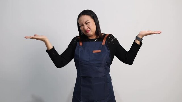 Adult asian female barista wearing a navy apron making a confused expression, raising both hands in a shrug gesture. Studio shot with white background, representing doubt, uncertainty, or not knowing