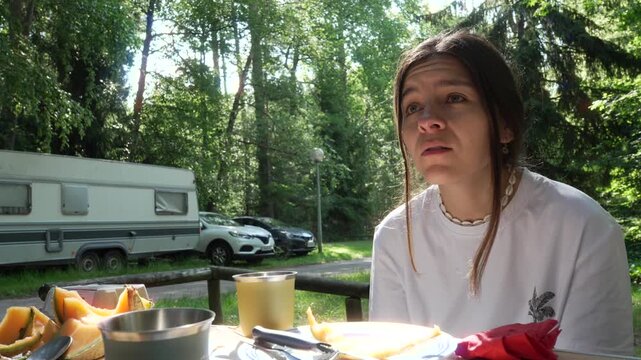A candid moment of a young woman enjoying breakfast and conversation at a sunny campsite.