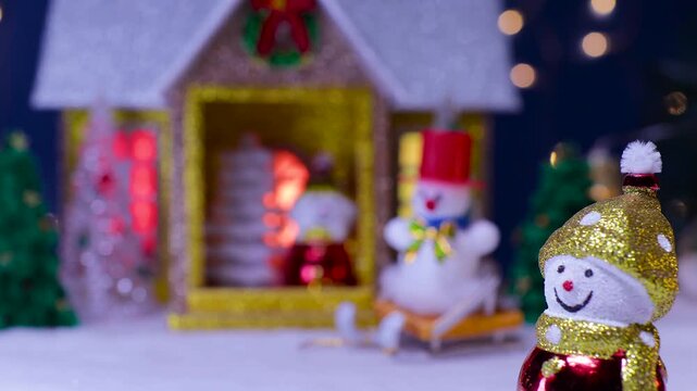 In front of the house with a snowman on the porch and a snowman on a sled nearby stands a small snowman