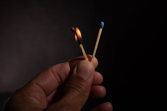 Hand holding two matches, one burning with a bright flame against a dark background