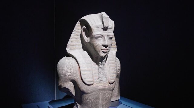 Stone bust of Ramesses II displayed in a dark museum setting, highlighting detailed nemes headdress and carved inscriptions under dramatic lighting.