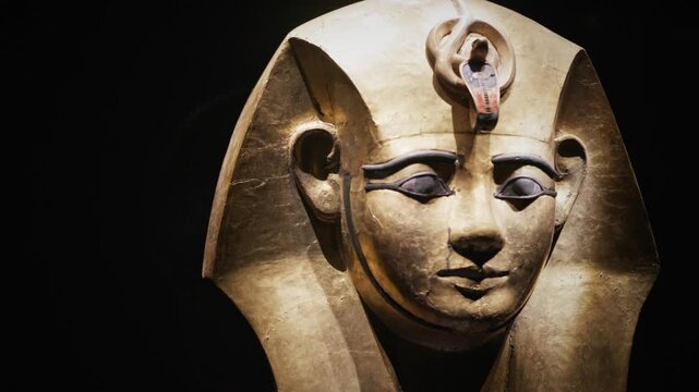 Close-up of a gilded ancient Egyptian coffin mask with detailed facial features and cobra ornament, displayed under museum lighting against a dark background.