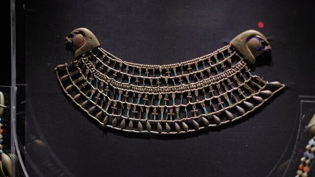 Close-up of an ancient Egyptian broad collar necklace exhibited in a museum display case, featuring intricate beadwork, rows of symbolic ankh motifs, and stylized falcon head terminals.
