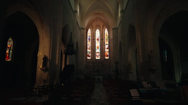 Dark interior of a religious building revealing colorful stained glass windows and a stone vault in Issigeac. Slow motion tracking shot.