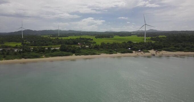 Aerial drone slowly descending over calm sea toward tropical coastline with three wind turbines generating renewable energy in Guimaras Philippines