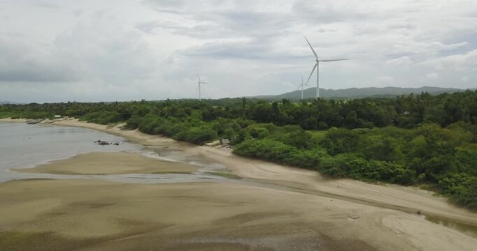 Static aerial view of tropical coastline with sandy beach and calm water while wind turbines rotate in the background generating renewable energy in Guimaras Philippines