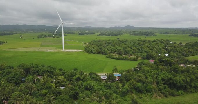 Aerial ascending shot of wind turbine rotating over lush tropical landscape with rural fields and vegetation in Guimaras Philippines under cloudy sky