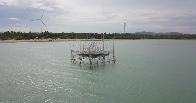 Aerial drone performing 360 orbit around traditional bamboo fishing platform over calm water with wind turbines along tropical coastline in Guimaras Philippines