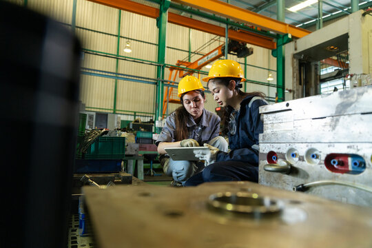 Female engineers inspecting metal mold parts in factory workshop, wearing safety helmets and gloves, checking industrial machinery with clipboard and tablet for quality control in manufacturing plant.
