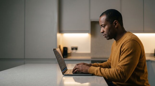 African American man coding on laptop while working in modern home kitchen.