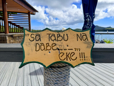 Hand carved wooden sign in Fijian language saying &ldquo;Sa tabu na dabe eke&rdquo; meaning do not sit here on tropical resort deck in Fiji with ocean and mountains in background
