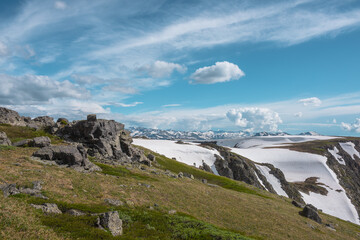 Scenic view to sunlit grassy hillside with rocky outcrops against snowfields on rocks and big snowbound rock mountain range far away under clouds in blue sky. High snow mountains in changeable weather © Daniil