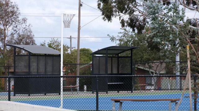 Fenced outdoor netball court with player shelters and seating in a suburban park in Australia. Community sports facility featuring court surface, hoop and surrounding traffic in a neighborhood