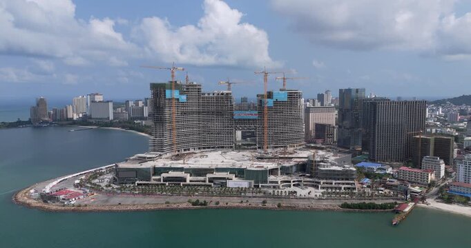 Ascending backward aerial view of massive coastal skyscrapers under construction in Sihanoukville, featuring numerous cranes and a modern urban skyline beside a sandy beach.