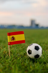 Spain national flag placed next to a soccer ball on a lush pitch during golden hour. Blurred stadium background adds a professional atmosphere, ideal for sports marketing and global football themes. © The Dell Shot by SJ