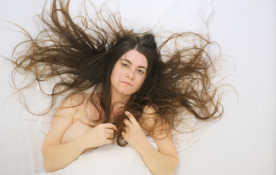 Young woman lying on white bed with long flowing hair spread around head looking calmly at camera expressing intimacy vulnerability softness rest and quiet emotional openness
