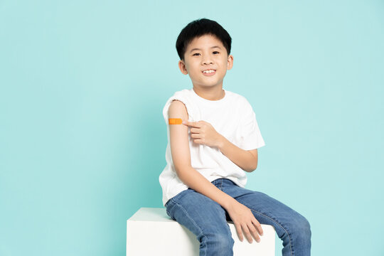 A smiling young Asian boy confidently sitting and shows off a bandage on his upper arm after receiving a vaccination isolated on green background. Pediatric healthcare, immunization campaigns