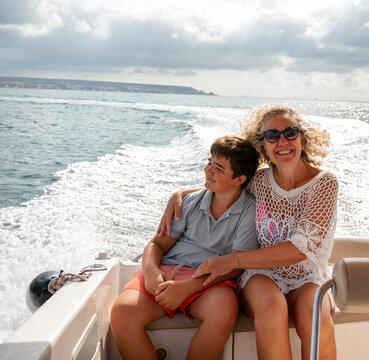 Middle aged woman embracing her teenager son on a motorboat during summer vacation, smiling on open sea with sparkling wake, sunshine, and candid family travel lifestyle
