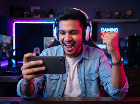 indian gamer holding smartphone and celebrating win with fist gesture in neon gaming environment for mobile gaming and esports concept