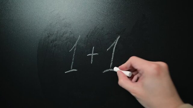 A hand writes a math equation on a blackboard using chalk. The action takes place in a classroom environment where simple arithmetic is taught