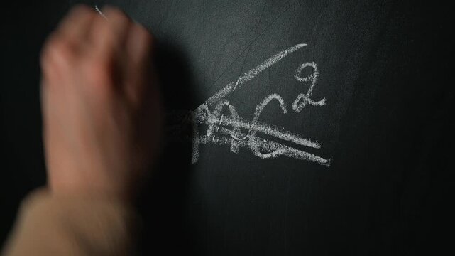 A hand appears on a chalkboard, initially writing the equation Emc, then crossing out each part repeatedly