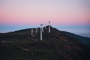 A group of white wind turbines stands on a mountain ridge above a thick cloud layer under a pink and purple twilight sky in a rural landscape during sunset. © JULIA