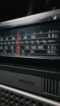 Close-up vertical view of an illuminated vintage analog radio tuner dial. A red indicator line sits over the frequency numbers. The retro receiver panel rests in a dark, moody environment.