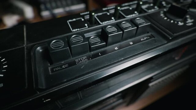 A person finger presses down a control button on a vintage black cassette tape recorder. The analog audio equipment features visible pause and stop mechanisms in a dimly lit setting.