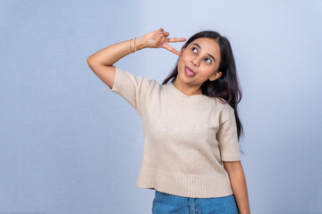 Funny Asian woman in beige shirt making peace sign and sticking tongue out on grey background © Jatmiko