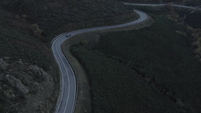 Mountain road in Serra da Estrela Seia municipality Guarda district Centro region Portugal showing car driving through S curve bordered by dense shrub vegetation, drone tracking descent shot
