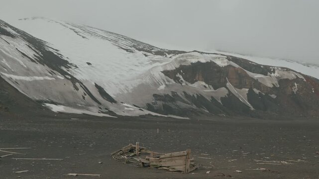 Decayed wooden whaling boat wreck lies on dark volcanic ash plain at Whalers Bay, Deception Island, dwarfed by sweeping snow-streaked rust-red volcanic slopes fading into pale mist