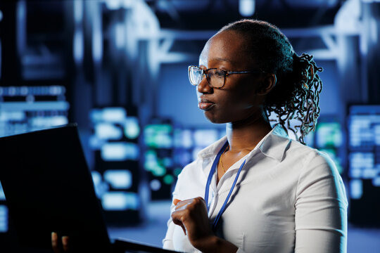 Proficient admin in high tech establishment uses laptop to prevent system overload. Competent expert between server cabinets ensuring enough network bandwidth for smooth operations