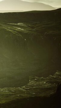 A landscape displays hills and cliffs with green vegetation under the sunlight. The scene shows natural terrain, creating a view of undulating hills and rocky formations.