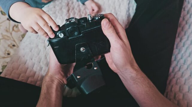 An overhead close-up shot of an adult hands holding a vintage black film camera. A small child hands reach out curiously to touch and explore the camera dials and buttons.