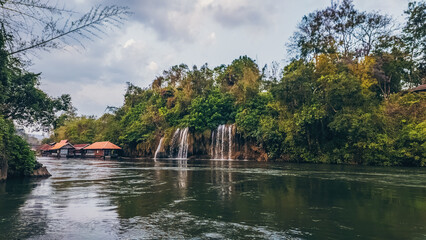Waterfall on river in Sai Yok Thailand with jungle landscape and reflections, Tropical cascade and floating houses along scenic riverbank in national park nature © goami